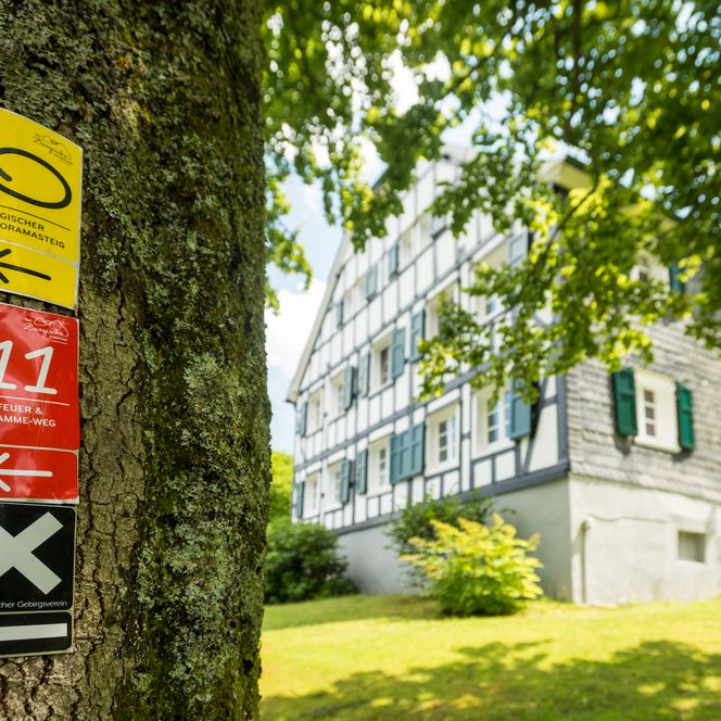 Ein Fachwerkhaus im Grünen, mit Wanderwegweisern an einem Baum im Vordergrund.
