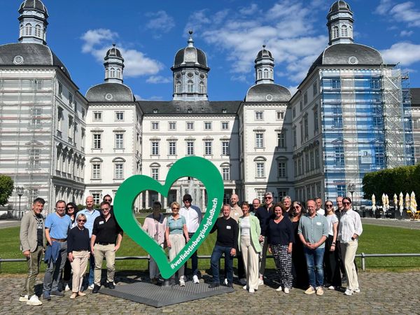 Die Teilnehmer einer Veranstaltung stehen bei schönem Wetter vor einem weißen Schloss. Vor ihnen steht ein großes grünes Herz.