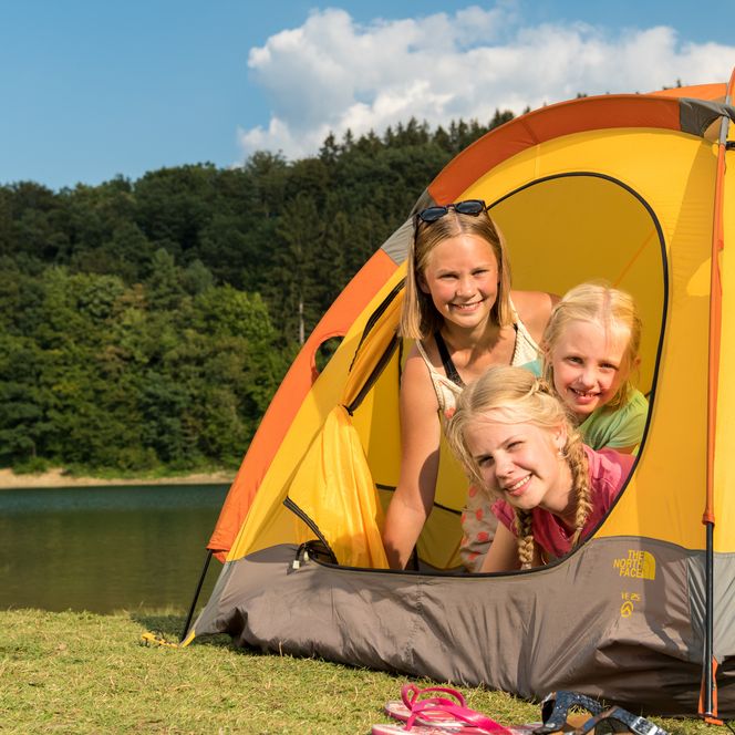 Eine Mutter mit zwei Kindern lacht im Zelt am Seeufer, umgeben von Wald und blauem Himmel.