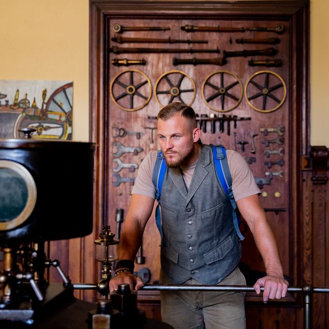 Mann betrachtet antike Maschinen in historischer Ausstellung, umgeben von Holzwänden und Werkzeugen.