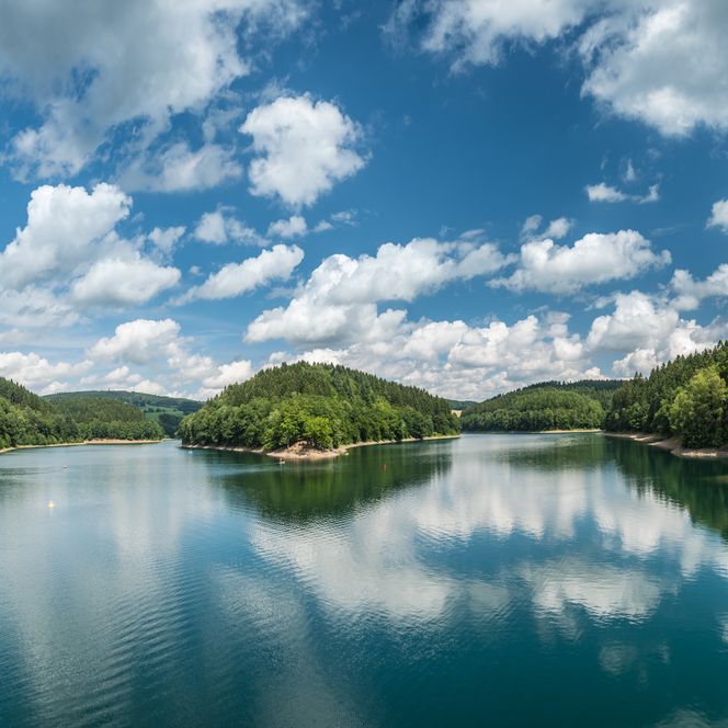 Panorama eines Sees in Gummersbach, umgeben von bewaldeten Hügeln und reflektierenden Wolken im Wasser.