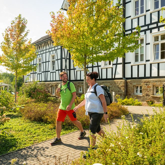 Fachwerkhaus in Much, Wanderer auf Gehweg, sonniger Herbsttag, blühende Büsche und Bäume.