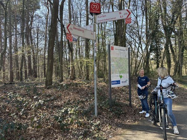 Zwei Radfahrerinnen stehen vor einer Infotafel im Wald. Eine schiebt ein Fahrrad, die andere hält einen Helm und eine Trinkflasche in den Händen.